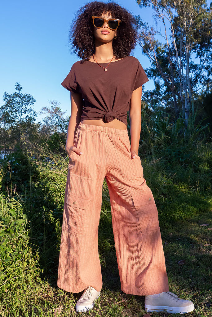 The Traveller Desert Pants are a gorgeous peachy orange pair of mid-rise cargo style pants. The pants feature an elasticated waist, side pockets, wide legs, and patch pockets. Made from 100% cotton.