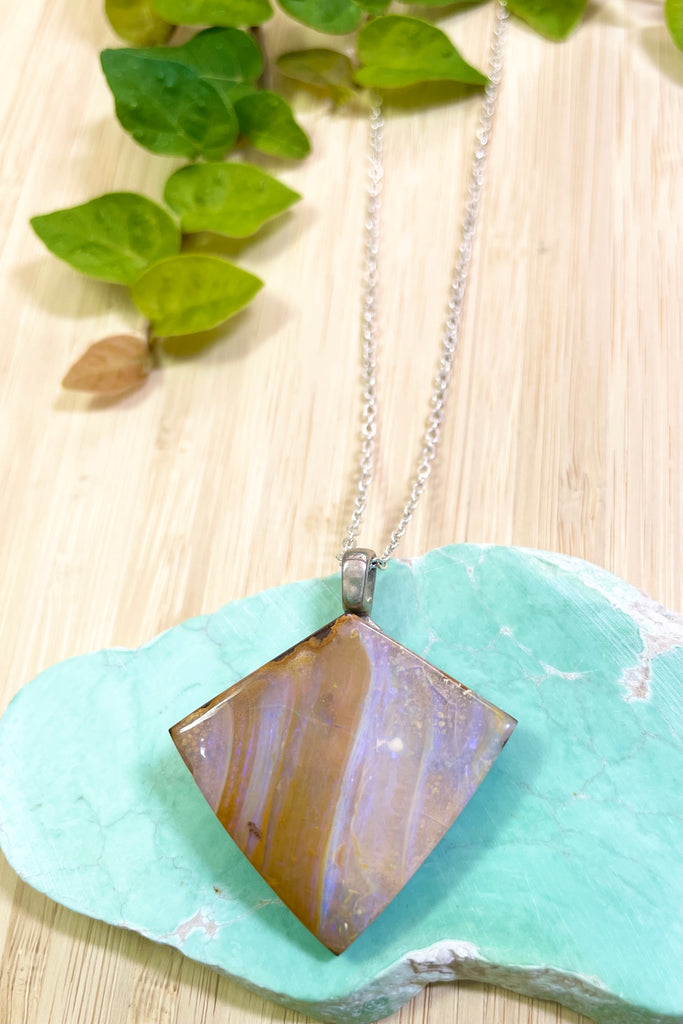 This opal is cut into a rough long triangle shape it has pale pinky blue opal flash with tiny flashes of green on the side. The face still shows some of the host rock from which it was cut.