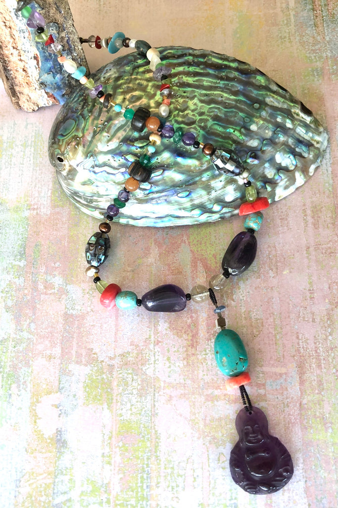 The elegant centre piece of this divine necklace is a mystical Deity carved in clear purple Amethyst and other stones on the chain include Peridot, Bad Howlite, Paua shell mosaic bead, green onyx, Carnelian, Amethyst, Golden Rutile Crystal and wood.