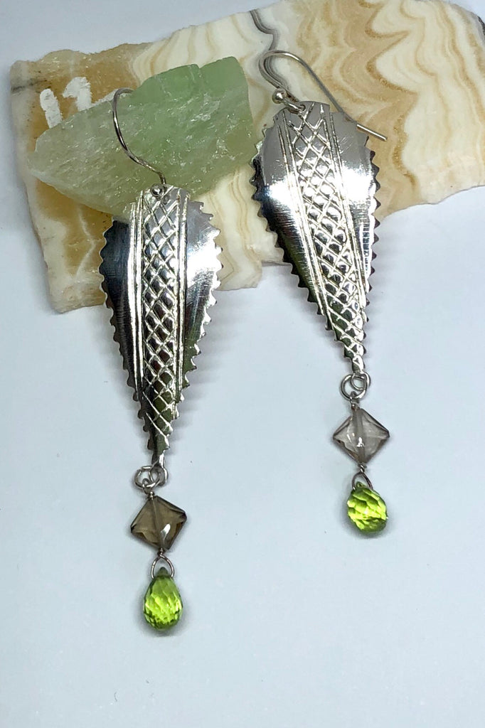 The African inspired design of these earrings was obtained by casting a pair of Mozambique Ebony wood earrings in silver. A Peridot gemstone briolet and a faceted smoky quartz hang from the base. 