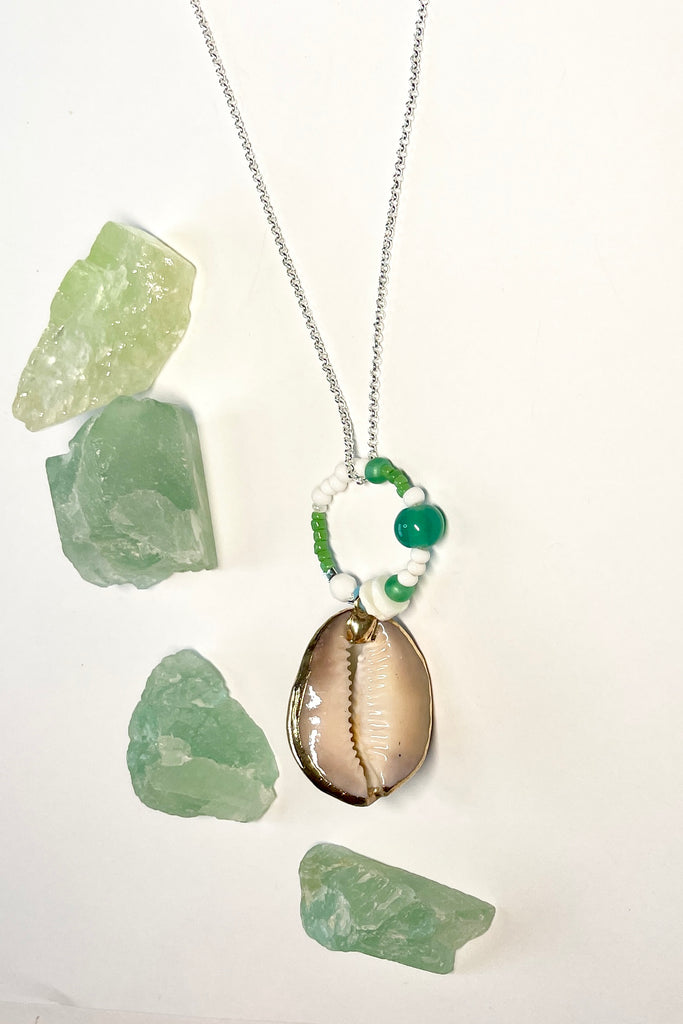 A pendant keepsake made with a cowrie shell which hangs from a circlet of simple green and white village beads. Then suspended from a chain.
