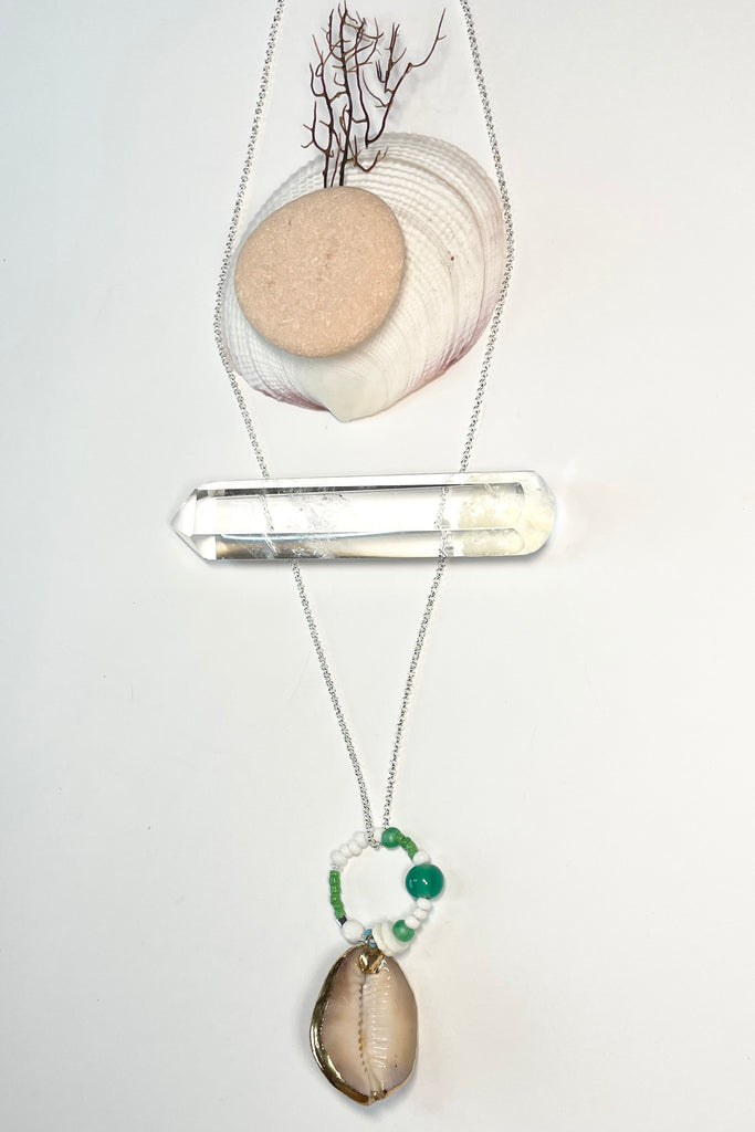 A pendant keepsake made with a cowrie shell which hangs from a circlet of simple green and white village beads. Then suspended from a chain.