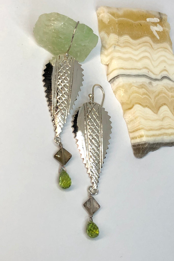 The African inspired design of these earrings was obtained by casting a pair of Mozambique Ebony wood earrings in silver. A Peridot gemstone briolet and a faceted smoky quartz hang from the base. 