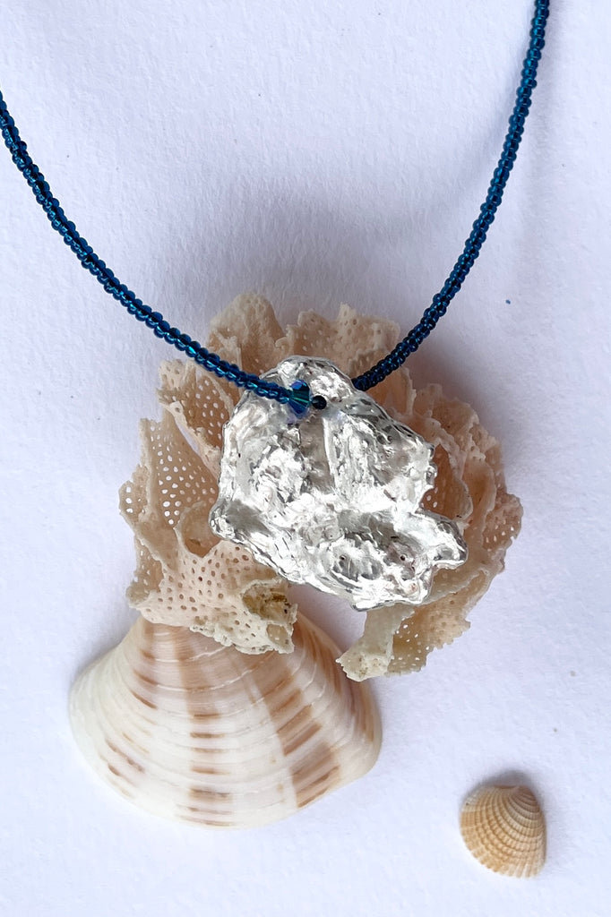 The centre piece is silver, it is cast from a shard of shell picked up on the Noosa beach. This organic shape of this pendant  is very unusual and unique. The underside is enameled in the colours of sea blue, with a scattering of sand and rocks, and bubbles. 