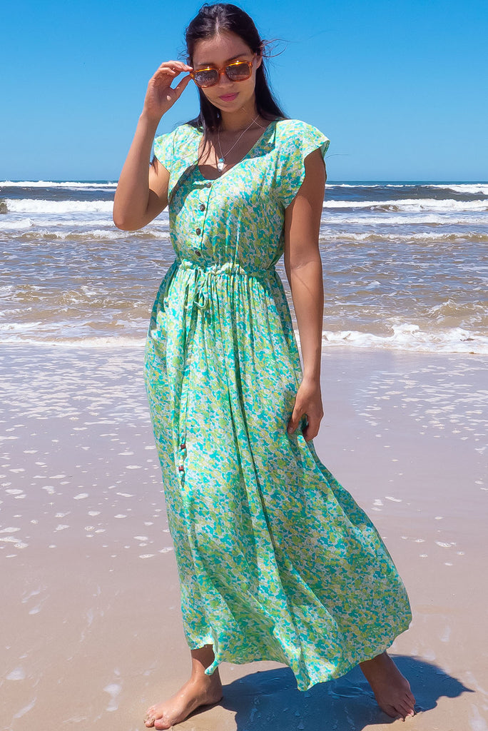 The Juniper Green Lemonade Maxi Dress is a vibrant green floral printed dress. The maxi dress features a functional button down chest to waistline, side pockets, adjustable drawstring waist, and cap sleeves. Made form woven 100% rayon.