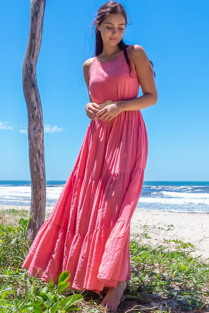 The Lulu Darling Coral Tiered Maxi Dress features a high neck with thin straps, high cut under arms, full tiered skirting from the bust down and deep side pockets. The dress is a coral pink colour made from a textured cotton and rayon blend. 