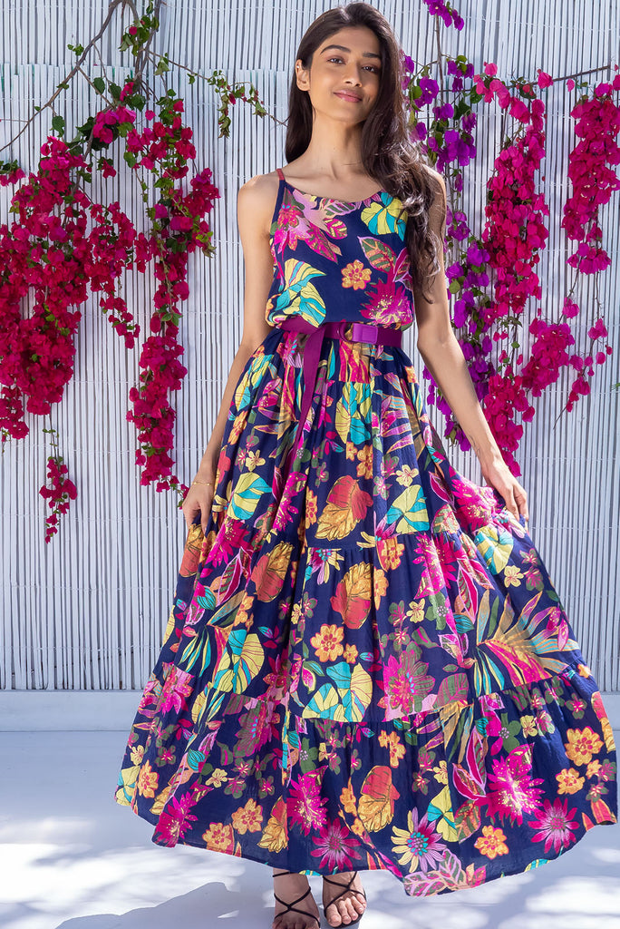 The Lulu Darling Paradise Navy Maxi Dress is a striking dress with an ink navy base and large tropical print. The maxi dress features a high neck, high cut under the arms, full tiered skirt falling from under bust, side pockets, and is woven cotton/rayon blend.