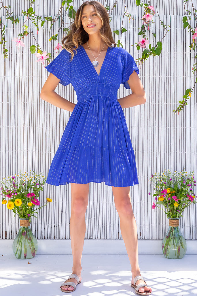 The Mirabella Harmonia Blue Dress is a beautiful blue mini dress with a silver lurex stripe. The dress features a lined bust, v neckline, drop flutter sleeves, low v back with tie at the top, cinched elastic waist, tiered skirt and side pockets. This dress is made from woven 100% crinkle rayon.
