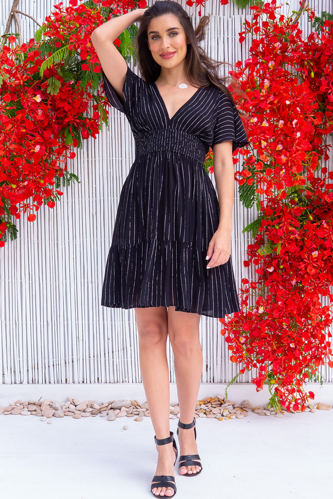 The Mirabella Noir Nights Dress is a stunning black mini dress with silver lurex stripes. The dress features a lined bust, v neckline, drop flutter sleeves, low v back with tie at the top, cinched elastic waist, tiered skirt and side pockets. This dress is made from woven crinkle rayon.