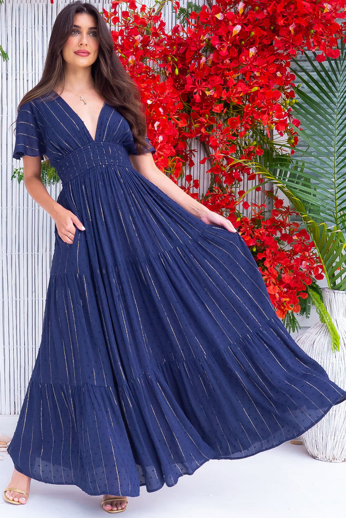 The Romance Navy Lurex Maxi Dress is a stunning navy maxi dress with textured spots and gold lurex stripes. The dress features a lined bust, deep plunge v neckline, drop flutter sleeves, low v back with tie at the top, cinched elastic was it, full tiered skirt and side pockets. This dress is made from woven crinkle rayon.
