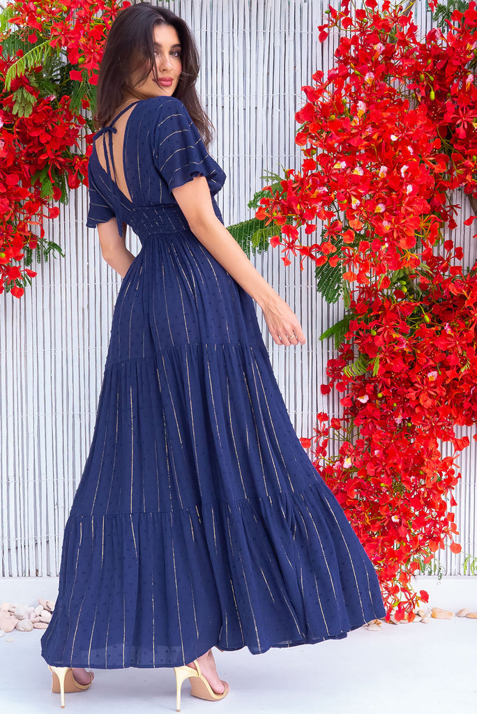The Romance Navy Lurex Maxi Dress is a stunning navy maxi dress with textured spots and gold lurex stripes. The dress features a lined bust, deep plunge v neckline, drop flutter sleeves, low v back with tie at the top, cinched elastic was it, full tiered skirt and side pockets. This dress is made from woven crinkle rayon.