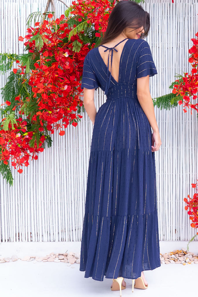 The Romance Navy Lurex Maxi Dress is a stunning navy maxi dress with textured spots and gold lurex stripes. The dress features a lined bust, deep plunge v neckline, drop flutter sleeves, low v back with tie at the top, cinched elastic was it, full tiered skirt and side pockets. This dress is made from woven crinkle rayon.