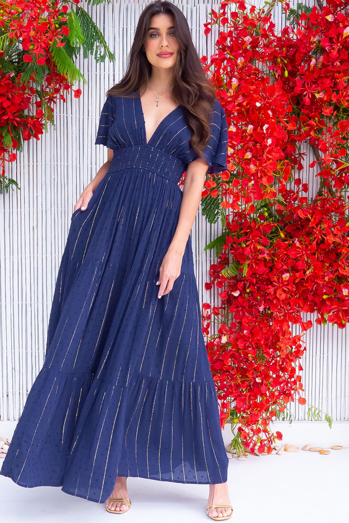 The Romance Navy Lurex Maxi Dress is a stunning navy maxi dress with textured spots and gold lurex stripes. The dress features a lined bust, deep plunge v neckline, drop flutter sleeves, low v back with tie at the top, cinched elastic was it, full tiered skirt and side pockets. This dress is made from woven crinkle rayon.