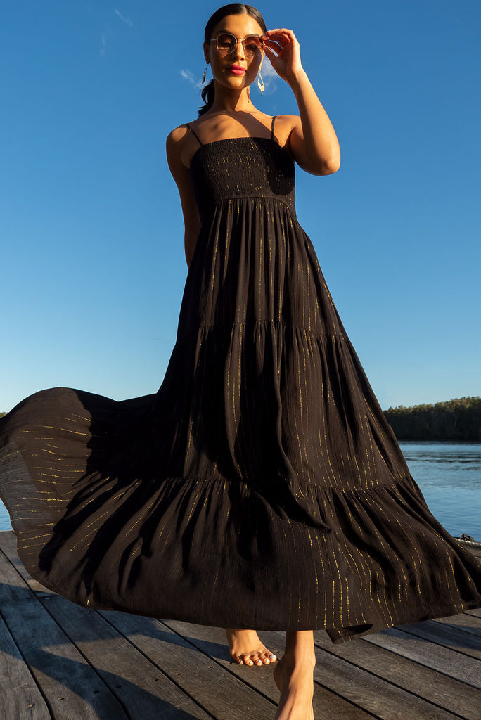 The Stardust Eclipse Noir Maxi Dress is a striking black dress with gold lurex stripes. This maxi dress features an elasticated shirred bodice, textured cheesecloth fabric, adjustable thin straps, tiered skirt and side pockets.