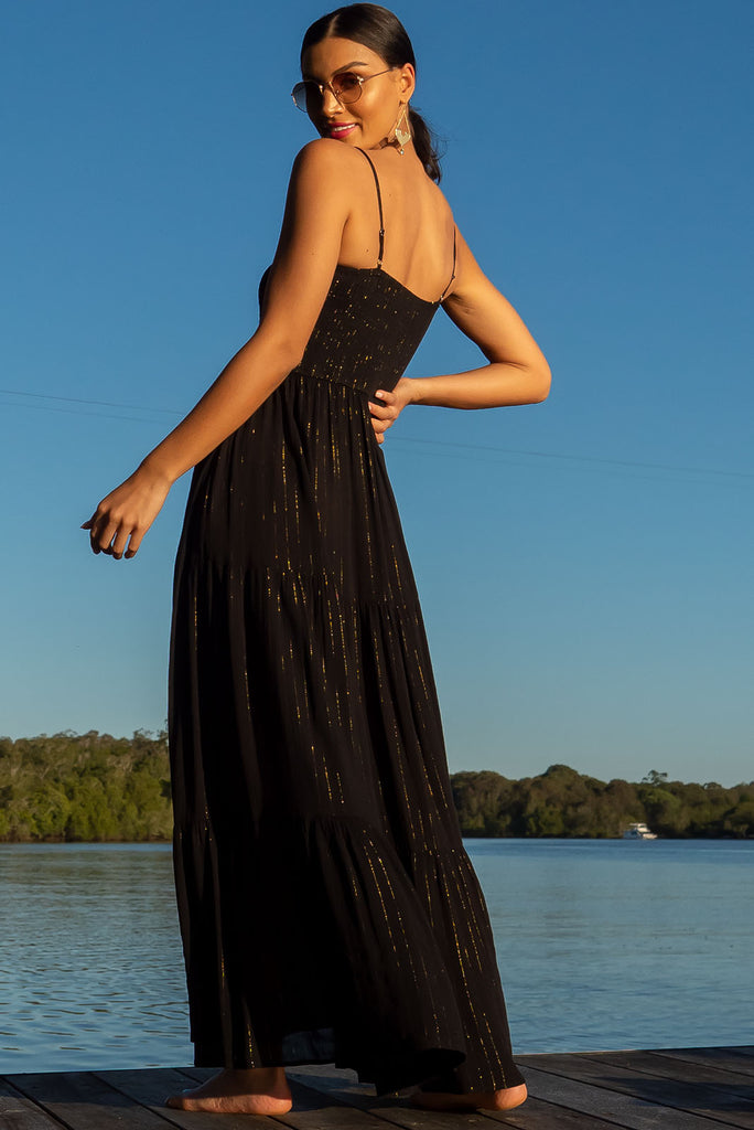 The Stardust Eclipse Noir Maxi Dress is a striking black dress with gold lurex stripes. This maxi dress features an elasticated shirred bodice, textured cheesecloth fabric, adjustable thin straps, tiered skirt and side pockets.