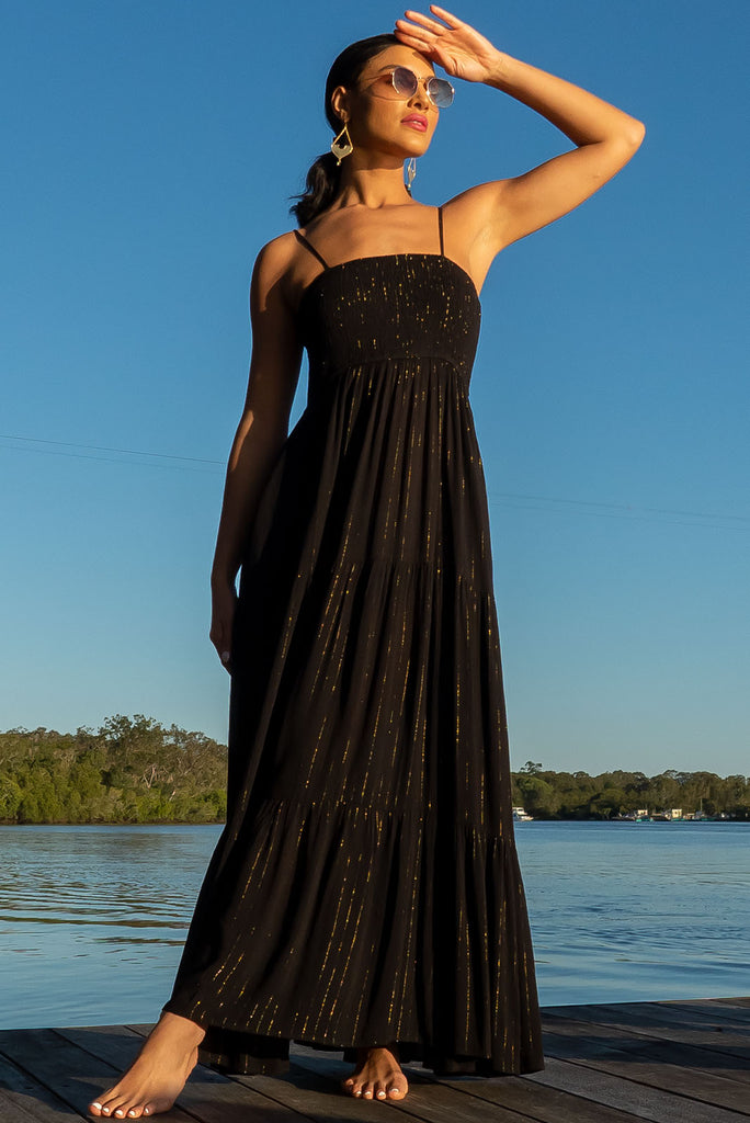 The Stardust Eclipse Noir Maxi Dress is a striking black dress with gold lurex stripes. This maxi dress features an elasticated shirred bodice, textured cheesecloth fabric, adjustable thin straps, tiered skirt and side pockets.