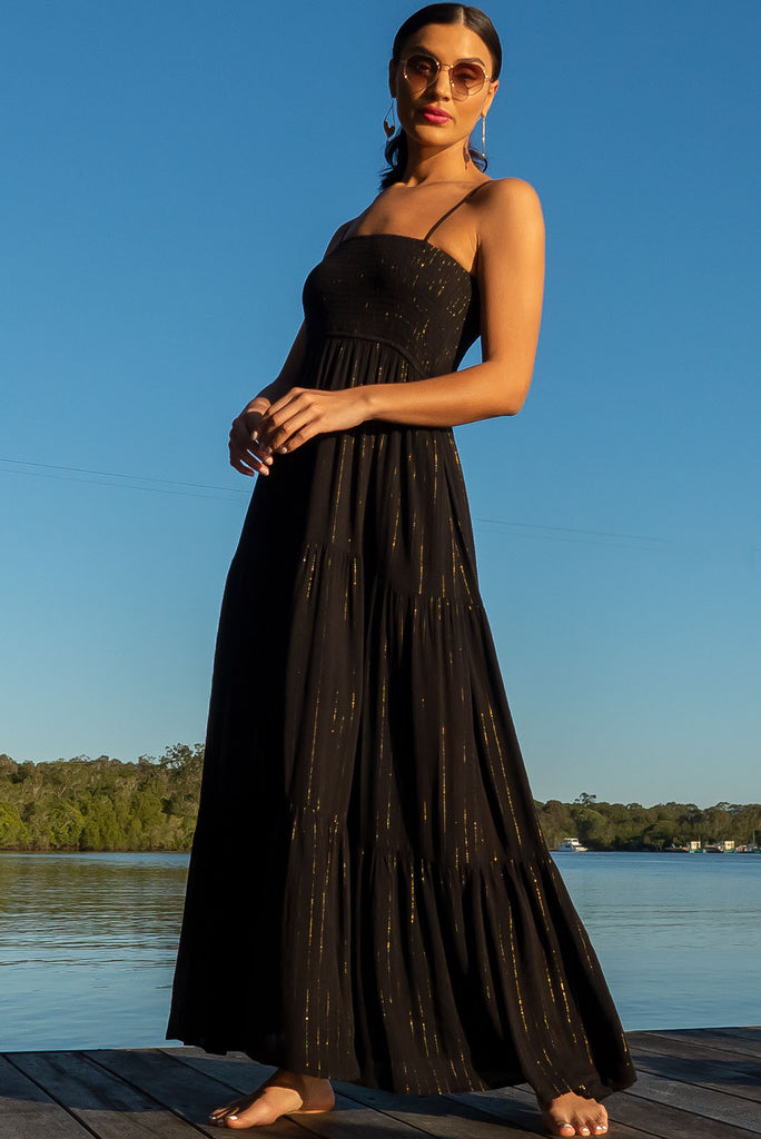The Stardust Eclipse Noir Maxi Dress is a striking black dress with gold lurex stripes. This maxi dress features an elasticated shirred bodice, textured cheesecloth fabric, adjustable thin straps, tiered skirt and side pockets.