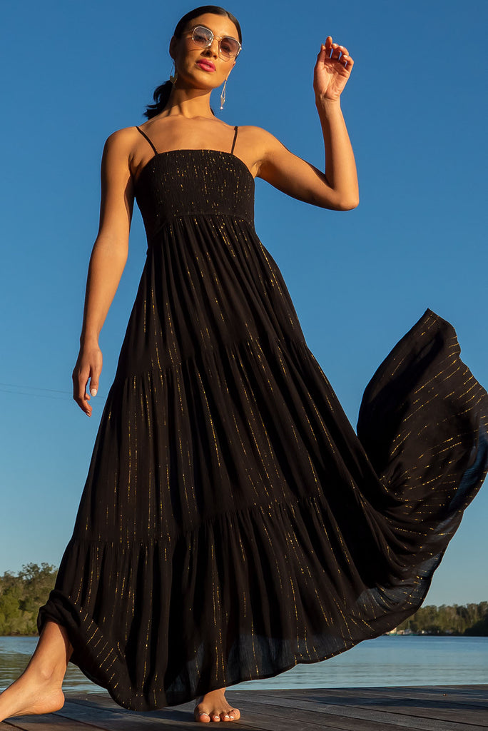 The Stardust Eclipse Noir Maxi Dress is a striking black dress with gold lurex stripes. This maxi dress features an elasticated shirred bodice, textured cheesecloth fabric, adjustable thin straps, tiered skirt and side pockets.